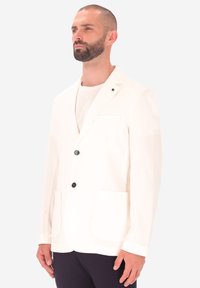 Uomo con barba corta che indossa un giubbotto bianco sopra una camicia bianca e pantaloni scuri, in piedi contro uno sfondo chiaro e semplice.