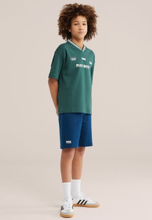 Mladost z zvitimi lasmi stoji oblečena v zeleno majico z napisom "BAD BOY", modre kratke hlače z napisom "SOCCER LEAGUE", bele nogavice in bele superge s črnimi črtami.