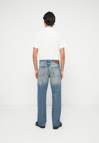 Man standing facing away wearing a white t-shirt, faded blue jeans, and black shoes against a plain white background.