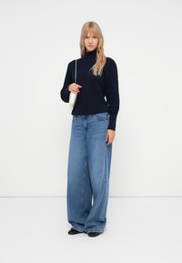 Mornarsko modra pulover s visokim ovratnikom in teksturiranim pletenjem, v kombinaciji z oversize svetloblue denim hlačami in črnimi čevlji. Na rami nosi majhno belo torbico.