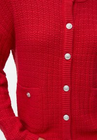 Gilet en tricot rouge avec un motif texturé, comportant des boutons en argent et une petite poche avant avec un détail boutonné.