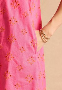 Rosa Stoff mit Blumenstickerei in orangefarbigen Tönen, ausgestattet mit einer Seitentasche und einer glatten Textur. Hervorgehobenes, einfaches, lässiges Design.