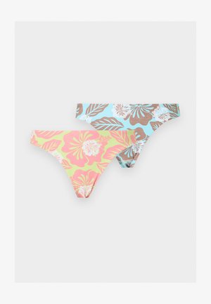 Jeune femme debout pieds nus, portant un maillot de bain deux pièces à motifs floraux pastel et des colliers superposés sur un fond clair uni.