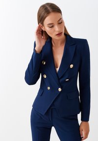 Blazer doppiopetto blu navy con bottoni dorati, rever a punta e tasche frontali. Tessuto liscio con vestibilità sartoriale, abbinato a pantaloni coordinati.