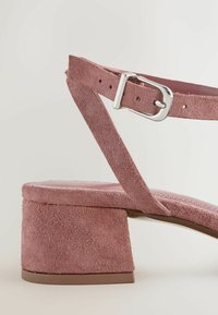 Sandale à talon bloc en daim rose avec bride à la cheville et boucle argentée, vue en gros plan sur un fond clair.