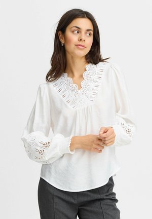 Vit blus med spetsdetaljer kring halsringningen och ärmsluten, med puffärmar och avslappnad passform. Texturerad tyg, lätt genomskinlig.