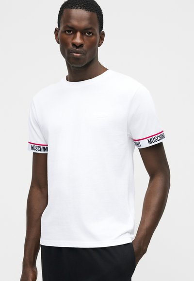 Άνδρας φοράει λευκό κοντομάνικο t-shirt με το κείμενο "MOSCHINO" στις μανσέτες των μανικιών και μαύρο παντελόνι, στέκεται μπροστά σε απλό ανοιχτό φόντο.