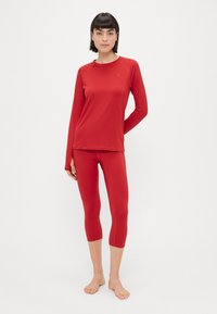Červený dlhý rukáv top a krátke legíny, oboje vyrobené z hladkého, elastického materiálu. Obsahuje okrúhly výstrih a jemný detail loga na vrchu.