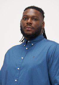 Modrá košeľa na gombíky vyrobená z hladkej tkaniny s jemným leskom. Má golier, predné gombíky a malé logo na hrudi.