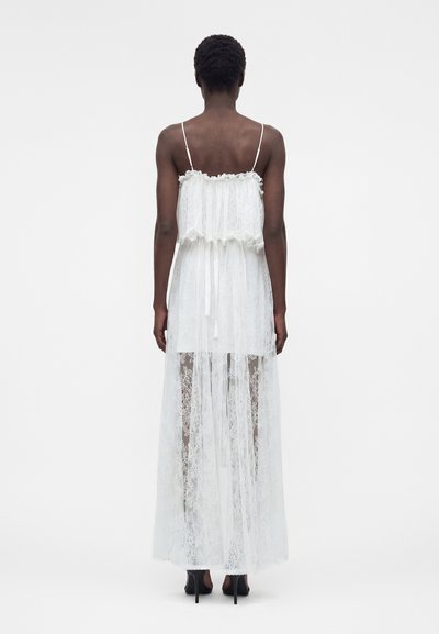 Γυναίκα με μακρύ διαφανές λευκό δαντελωτό φόρεμα με λεπτές τιράντες, δεμένη στη μέση, και μαύρα ψηλοτάκουνα παπούτσια, στέκεται με την πλάτη προς τα έξω σε απλο φόντο.