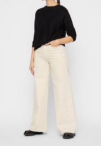 Pull noir à manches longues associé à un pantalon pattes d'éléphant beige clair. Le mannequin se tient les mains dans les poches, portant des chaussures noires. Design simple.