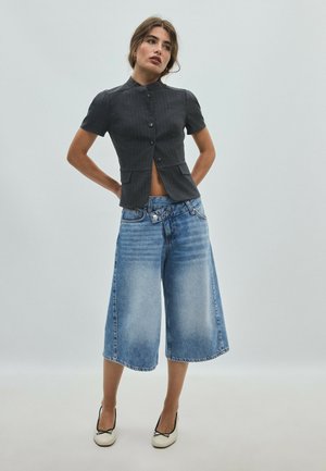 Woman wearing a short-sleeve black buttoned top and wide, knee-length blue denim pants with white shoes, standing with hands behind back.