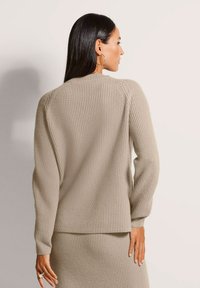 Frau mit langen dunklen Haaren, die einen beigen gerippten Strickpullover und einen passenden Rock trägt, mit dem Rücken zur Kamera, vor einem hellen Hintergrund.