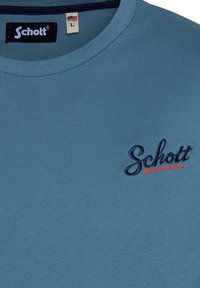 Camiseta de algodón azul claro con cuello redondo, que presenta un logo bordado de "Schott" en color navy y texto acentuado en rojo.