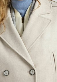 Beige double-breasted coat with wide lapels, two large gray buttons, and a textured fabric. Layered with a light blue turtleneck and checked inner lining.