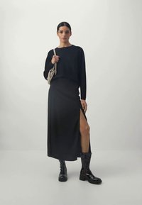 Schwarzes Langarmoberteil kombiniert mit einem schwarzen Rock mit hohem Schlitz. Das Model trägt glänzende schwarze Overknee-Stiefel und hält eine kleine beige Tasche.