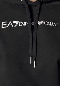 Svart huvtröja med EA7 Emporio Armani-logotyp i vitt. Inkluderar svarta dragsko med silverfärgade aglets. Mjuk tygtextur.