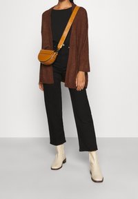 Brauner gestrickter Cardigan, schwarzes Oberteil, hochgeschnittene schwarze Hose, cremfärbige Knöchelstiefel und eine kleine senfgelbe Tasche mit einem Schultergurt.