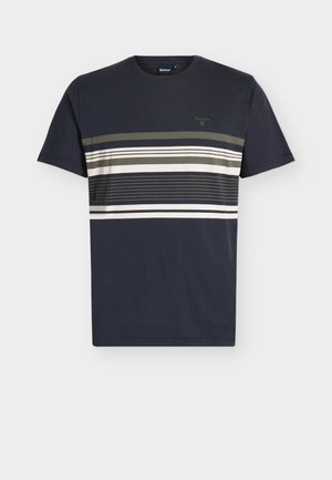 Homme en T-shirt rayé bleu marine foncé et pantalon beige tenant une veste beige assortie, portant des baskets blanches avec des touches bleues, regardant vers la droite.