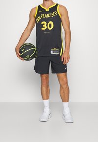 Mies jalkapalloilija, joka käyttää mustaa ja keltaista San Franciscon pelipaitaa numero 30, pitää mustaa ja keltaista koripalloa kädessään ja hänellä on valkoiset lenkkarit.