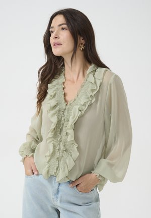 Blouse vert clair avec des volants le long de l'encolure et sur le devant, fabriquée en tissu transparent. Présente des manches longues bouffantes avec des poignets élastiques.