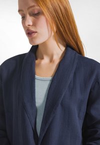 Granatowa lniana marynarka z kołnierzem typu szal, w połączeniu z jasnoniebieskim prążkowanym topem. Tkanina ma lekko teksturowane wykończenie i luźny krój.