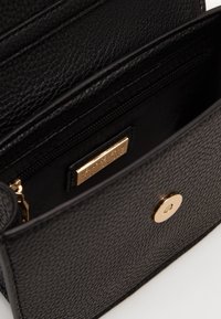 Svart texturerad läderväska öppen för att visa inre dragkedjefack med guldplatta och rund magnetlås på framfliken.