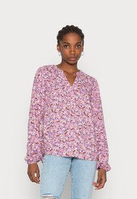 Blusa a maniche lunghe di colore viola chiaro con motivo floreale, dotata di scollatura a V e polsini elasticizzati. Abbinata a jeans chiari strappati.