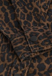 Gros plan d’un tissu marron avec un imprimé léopard noir et des boutons-pression noirs cousus sur les poignets du vêtement.