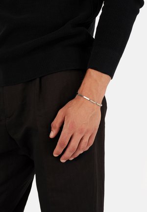 Silbernes Armband mit geometrischen Gliedern, das an einer Hand getragen wird. Das Handgelenk ist unbedeckt und zeigt den Hautton auf einem strukturierten schwarzen Pullover und einer Hose.