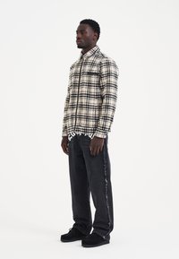 Camicia a quadri con bottoni in crema e nero, dotata di taschino sul petto. Pantaloni in denim neri con bordi sfrangiati. Scarpe nere. Design minimalista.