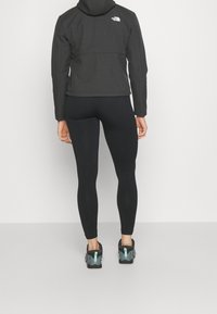 Musta hupullinen takki, istuvat mustat leggingsit ja kengät, joissa on sinivihreitä yksityiskohtia. Sileä kangas, minimalistinen design ja takissa näkyvä brändilogo.