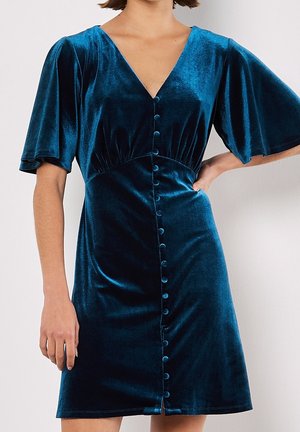 Kobieta ubrana w sukienkę z zielonkawoniebieskiego aksamitu z falującymi rękawami, dekoltem w serek i rzędem guzików obciągniętych tkaniną z przodu.