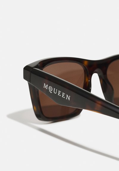 Occhiali da sole tartarugati con lenti marroni scure. Montatura spessa e angolare. L'asta sinistra riporta "MCQUEEN" in lettere bianche. Finitura lucida.