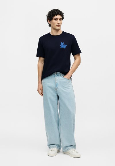 Άνδρας με μαύρο μπλουζάκι με μπλε εικόνα κουνελιού, ανοιχτό μπλε φαρδιά τζιν, λευκά αθλητικά παπούτσια, στέκεται με το ένα χέρι στην τσέπη σε λευκό φόντο.