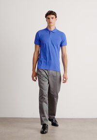 Jovem a caminhar, vestindo uma camisa polo azul, calças cinzentas e sapatos pretos de vestir, sobre um fundo branco liso.