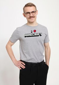 Šedé tričko s krátkým rukávem, na kterém je černý pixelový text "I ❤️ MINECRAFT" a malé pixelové srdce, nošené s černými kalhotami a opaskem.