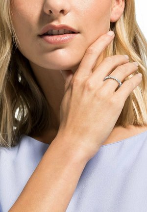 Frau mit blonden Haaren, die ein hellviolettes Oberteil trägt, berührt ihr Gesicht und zeigt einen silbernen Ring mit mehreren runden Diamanten an ihrem Finger.