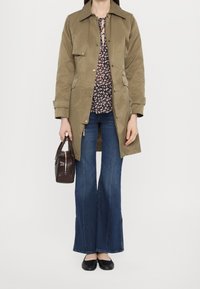 Trench-coat kaki porté sur un chemisier à fleurs, associé à un jean évasé et des ballerines noires. Un petit sac à main marron complète la tenue.