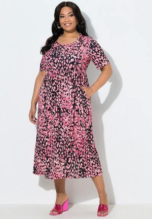 Donna che indossa un abito midi floreale rosa e nero con maniche corte, posa con una mano in tasca, indossando sandali rosa con tacco largo e punta aperta.