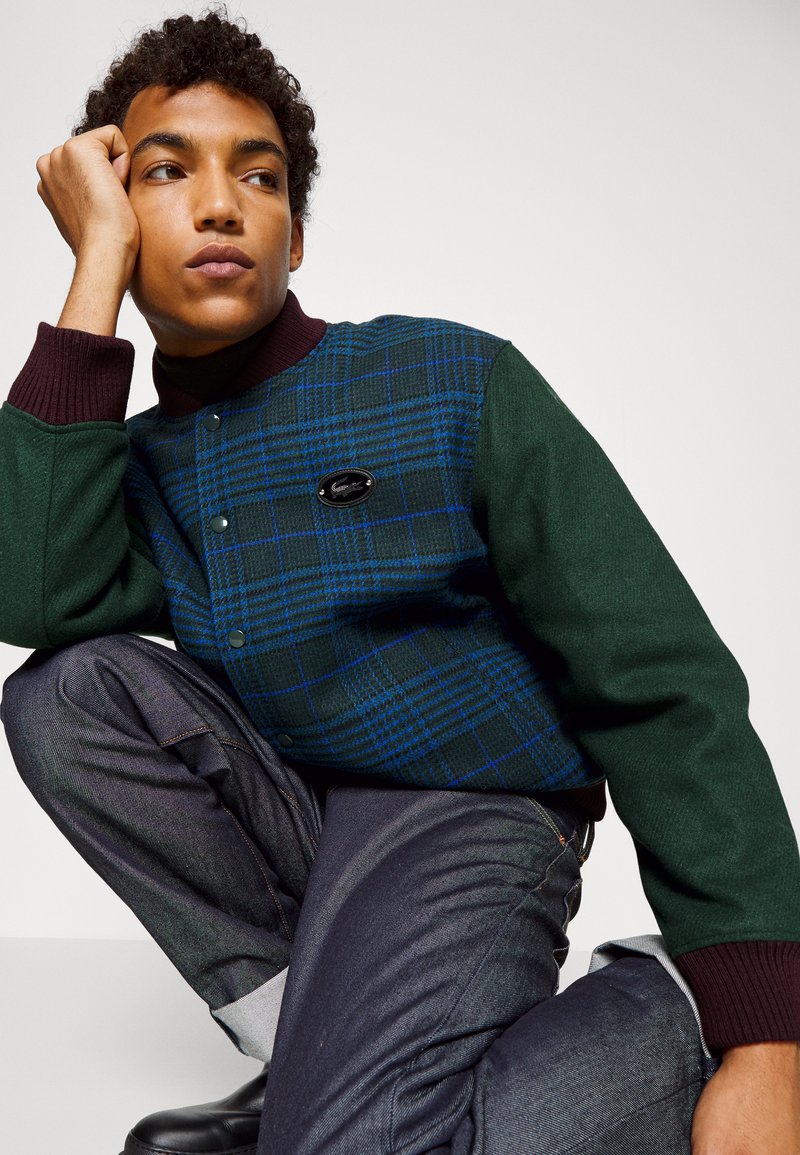 Veste bomber à carreaux en vert foncé et bleu, avec un col bordeaux et un écusson noir, associée à un pantalon en denim noir.