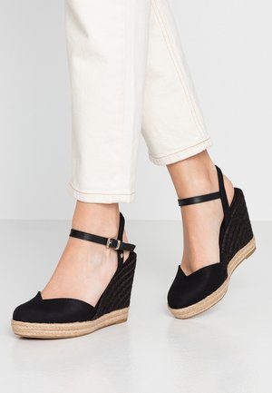 Pieds portant des sandales espadrilles compensées noires avec brides à la cheville et pantalon écru sur un fond clair.