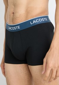 Boxers en coton noir avec une ceinture élastique bleue portant le logo "LACOSTE". Texture lisse et design ajusté.