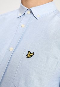 Camisa azul claro de manga corta con botones y un bolsillo, con un logo bordado en negro y dorado de un pájaro en el pecho.