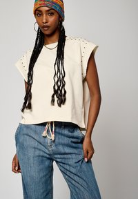 T-shirt crème cropped avec des accents d'épaules cloutés, associé à un pantalon en denim bleu ample avec taille à cordon et texture subtile.