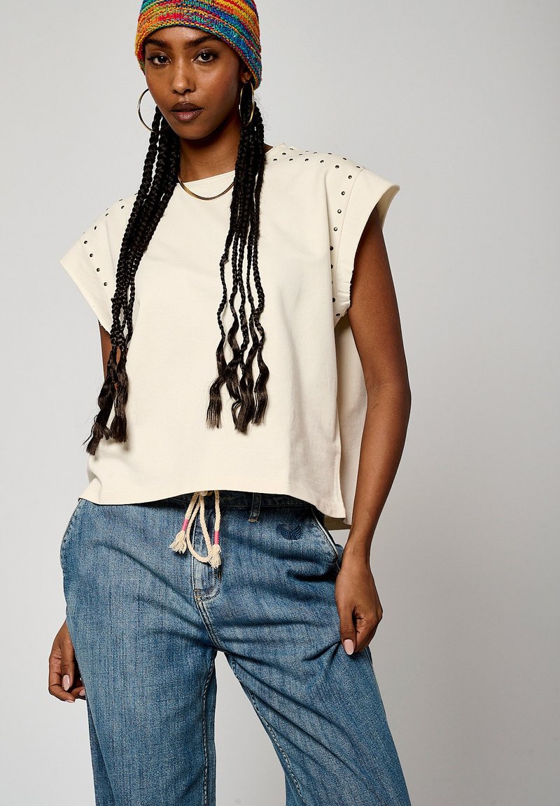 T-shirt crème cropped avec des accents d'épaules cloutés, associé à un pantalon en denim bleu ample avec taille à cordon et texture subtile.