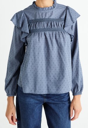 Blouse bleue à manches longues avec empiècement à volants, détails en dentelle, motif à petits pois subtil, portée avec un jean en denim bleu par une personne debout sur fond blanc.