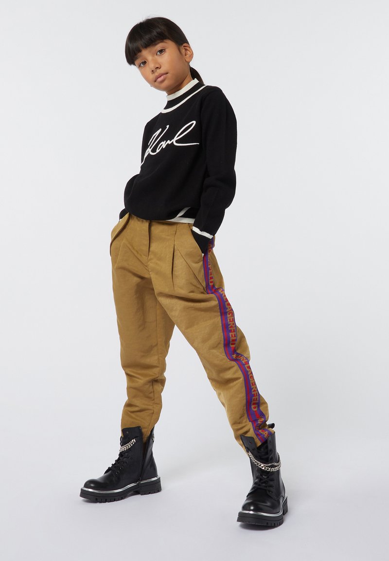 Zwarte trui met witte handtekeningontwerp, beige geplooide broek met paarse en rode zijstrepen en zwarte enkellaarzen met zilveren kettingdetail.