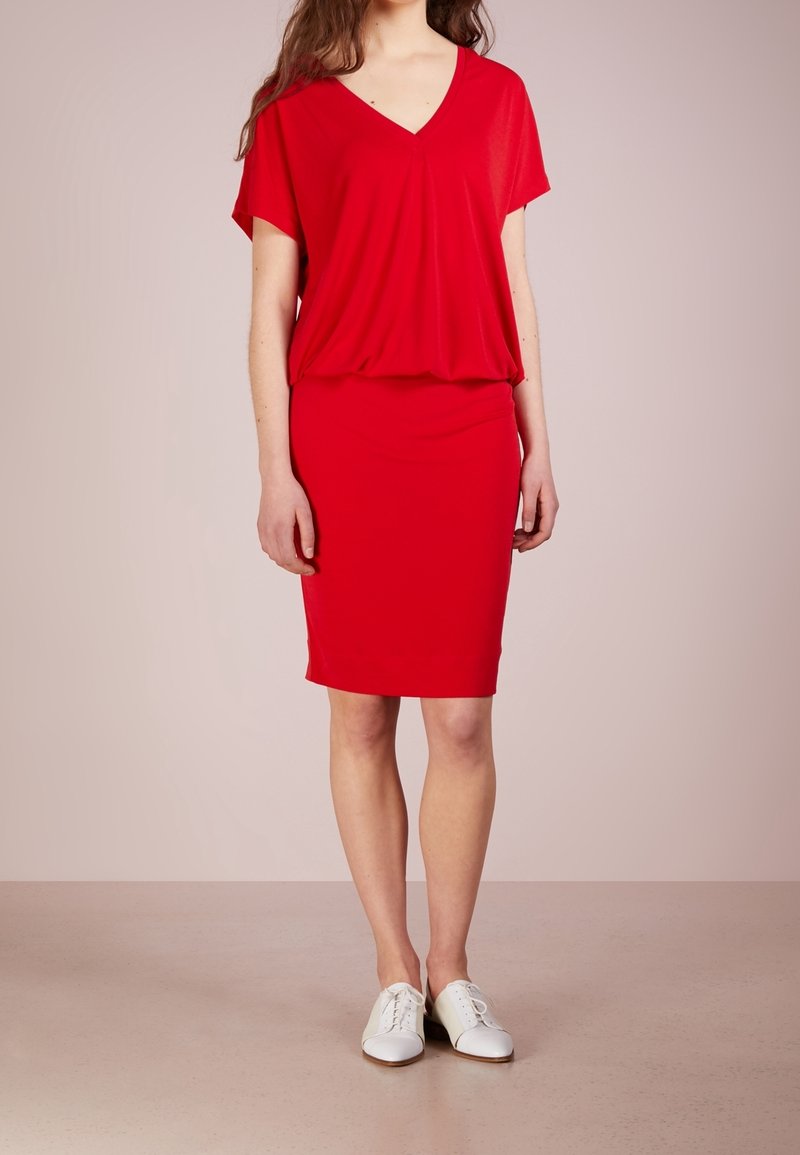 Femme portant une robe rouge à manches courtes avec un col en V, une jupe ajustée et des chaussures blanches à lacets, debout devant un fond uni.