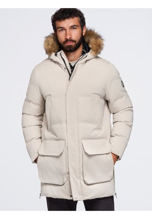 Homme portant un parka matelassé beige avec capuche bordée de fourrure et grandes poches avant, debout devant un fond clair uni.